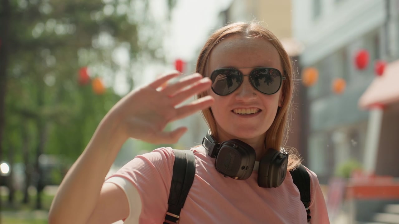 Una giovane donna con i capelli lunghi, che indossa una maglietta rosa e delle cuffie, cammina all'aperto in un ambiente urbano. La donna porta uno zaino e a un certo punto alza casualmente la mano per aggiustare le cuffie