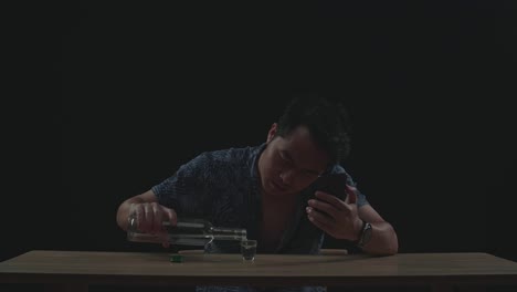 asian man pouring vodka in a shot glass before drinking during having video call in black background