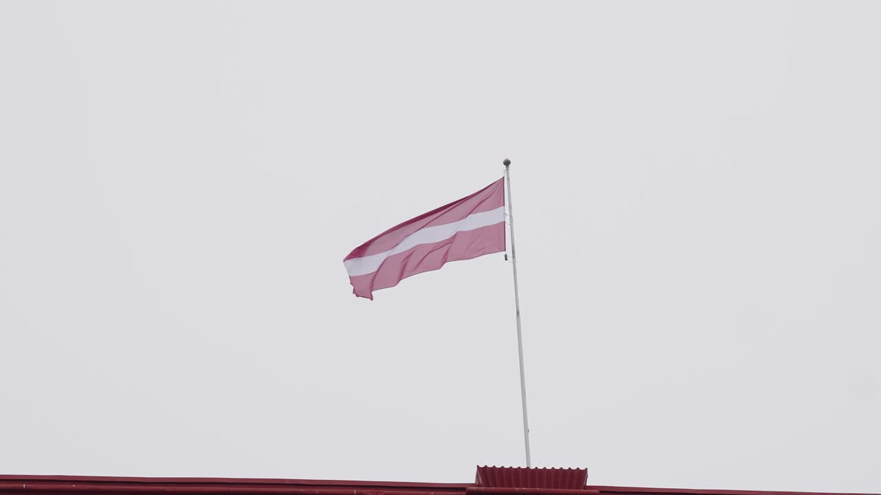 A Latvian national flag waves in the wind atop a building with a red roof.