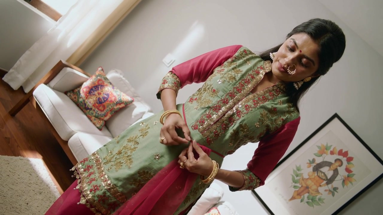Donna indiana che si sistema, allacciando una kurta verde e lisciando un dupatta rosa a casa con bracciali d'oro