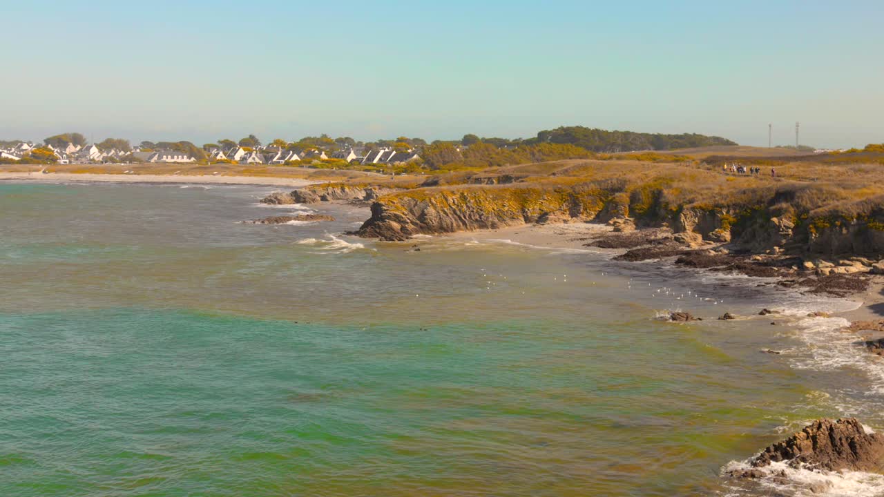 La costa frastagliata e rocciosa della Côte Sauvage a Quiberon, in Francia, che mostra il mare turchese in contrasto con la terra costiera preservata e un gruppo di case con il tetto bianco in lontananza