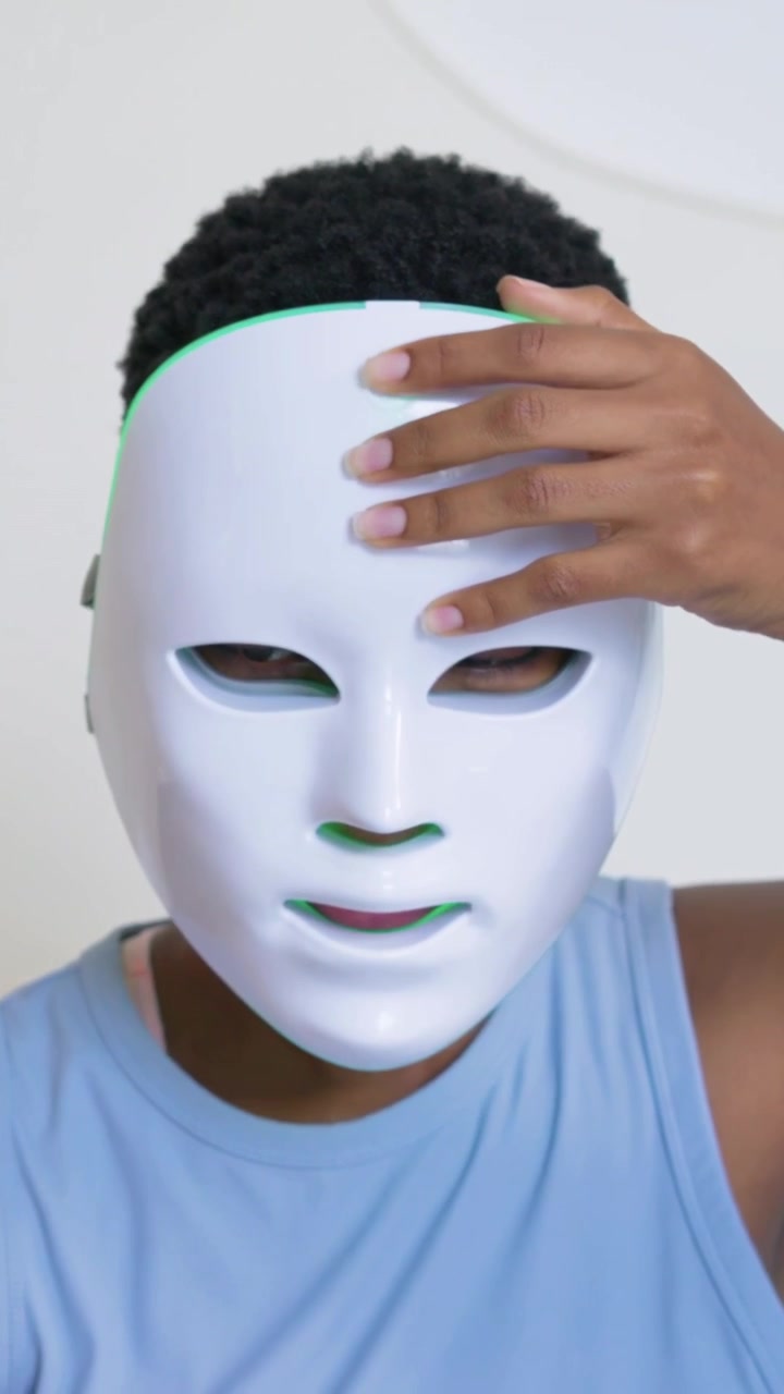 Vertikales Video: Frau hält LED-Maske an der Stirn, positioniert und drückt sie an für Hautpflege zu Hause, blaues Oberteil.