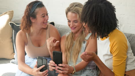 on couch, diverse female friends laughing and sharing smartphone moments, having fun, at home
