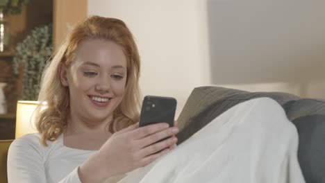 Mujer-Sonriente-En-Casa-Relajándose-En-El-Sofá-Mirando-El-Teléfono-Móvil