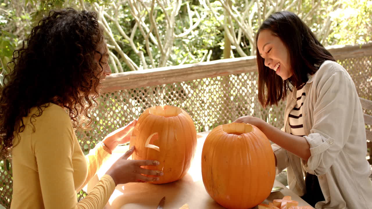 Halloween time, two multiracial female friends carving pumpkins on a porch