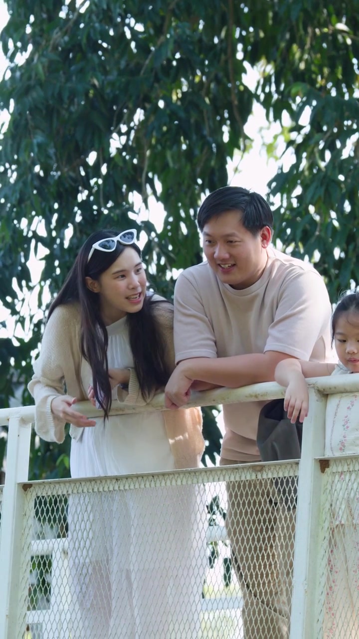 พ่อแม่และลูกสาวตัวน้อยกำลังพิงราวสะพานสีขาวในวันที่มีแดดจ้า
