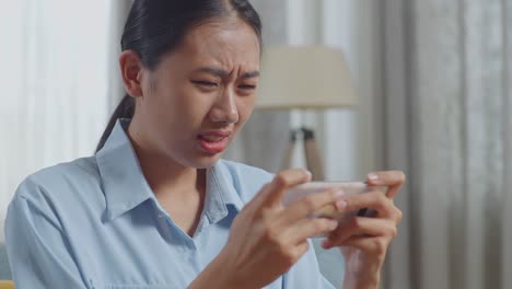 거실의 소파에 앉아있는 동안 스마트폰에서 게임을 잃는 아시아 여성의 가까운 모습