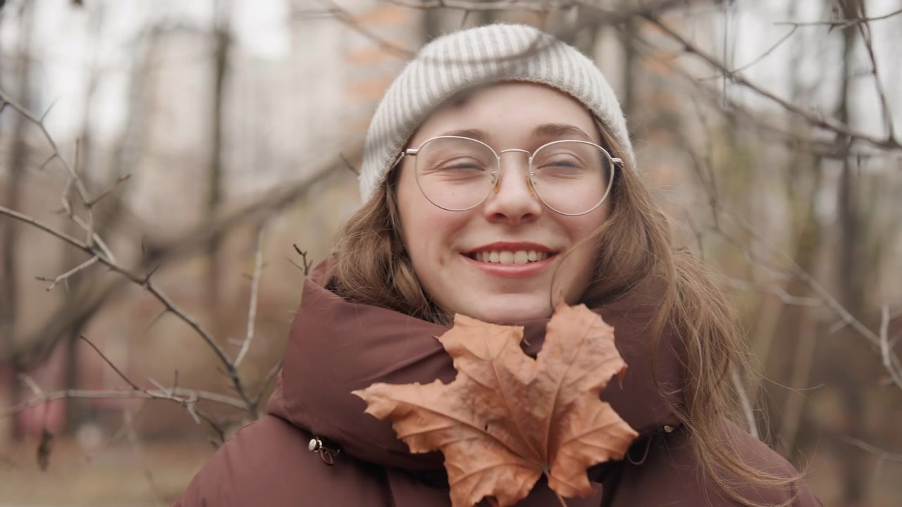 Giovane donna con berretto che tiene una foglia, sfondo di un parco autunnale delicato con alberi spogli e tonalità tenui, occhiali e sciarpa calda creano un ritratto accogliente, sorriso gentile e sguardo pensieroso, atmosfera intima all'aperto