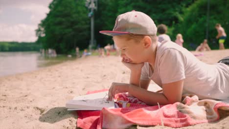 Süßer-Kleiner-Junge-Mit-Einer-Mütze,-Der-An-Einem-Sandigen-Seeufer-Liegt,-Das-Buch-Liest-Und-Seinen-Tropischen-Urlaub-Hat