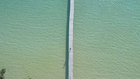 Aerial,-top-down,-birdseye,-drone-shot-over-a-woman-walking-on-a-wooden-pier-and-people-swimming-in-the-turquoise-sea,-at-a-paradise-beach,-on-a-sunny-day,-in-Koh-Kood,-Thailand,-Asia