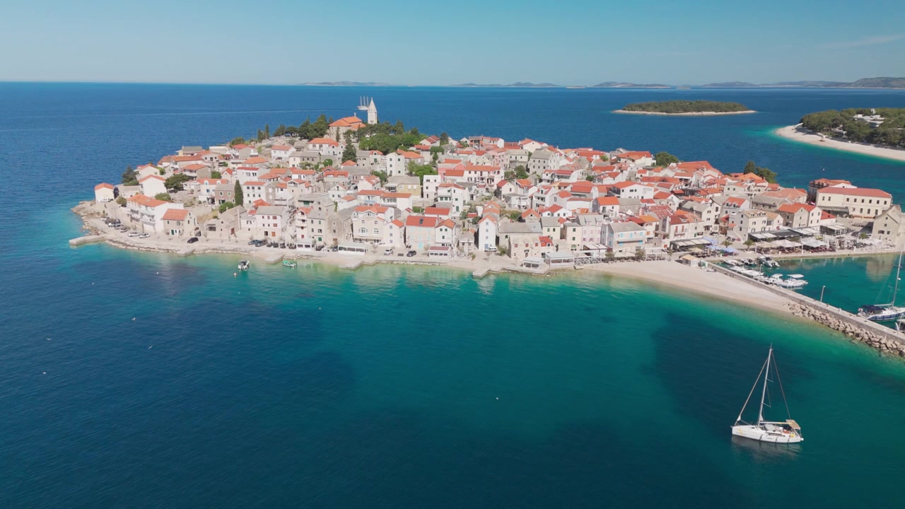 Ripresa laterale con drone sopra l'ex passaggio marittimo di Primošten e il piccolo molo per yacht, mantenendo una vista costante sulla penisola del centro storico e sul campanile della chiesa sopra l'Adriatico.