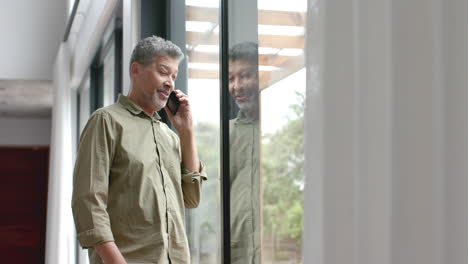 Feliz-Hombre-Birracial-Mayor-Hablando-En-Un-Teléfono-Inteligente-Parado-Junto-A-La-Ventana-En-Casa,-Espacio-Para-Copiar,-Cámara-Lenta