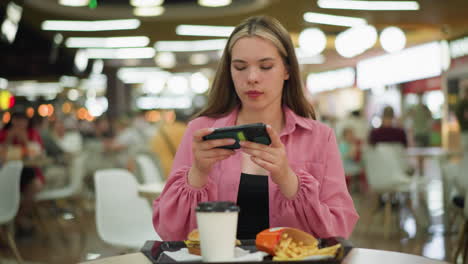 dama blanca en vestido rosa sentada en una mesa de madera en un restaurante ocupado, ajustándose para tomar una foto de su comida, la comida consiste en una hamburguesa, papas fritas y una taza de café en una bandeja negra