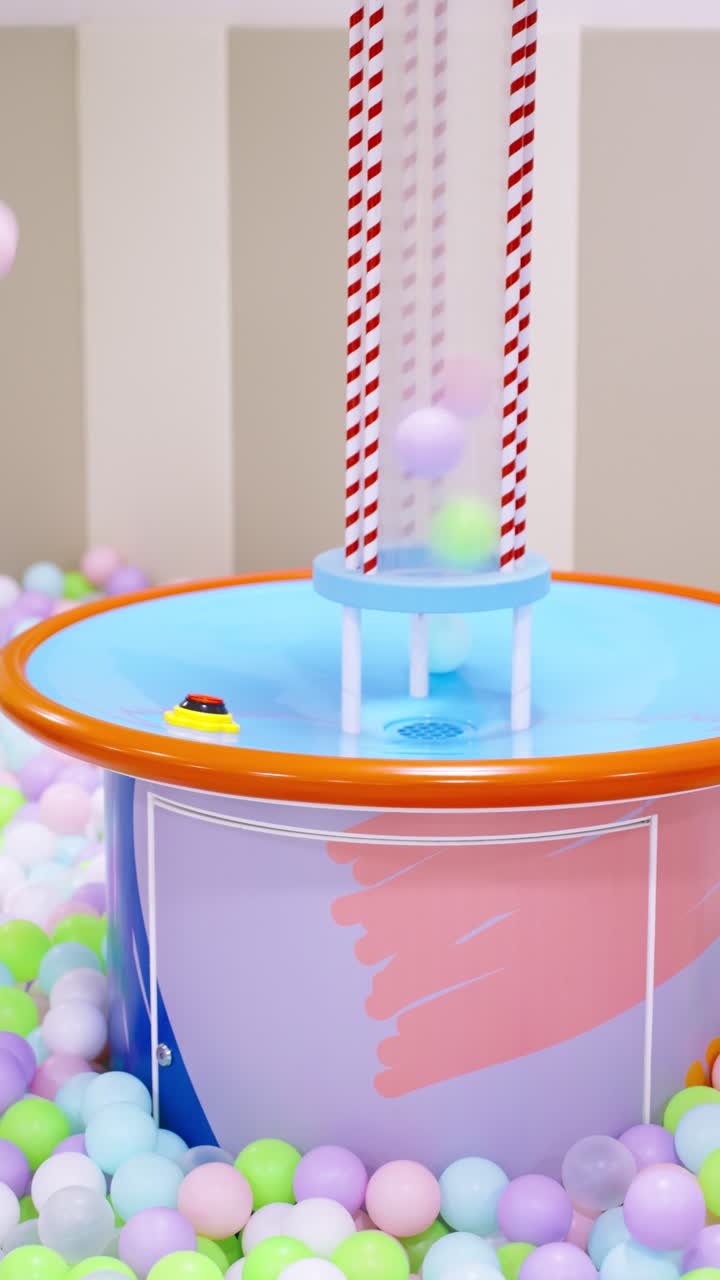 Little girl playing in a ball pit with an interactive ball-blowing machine, surrounded by colorful pastel balls - slow motion, vertical