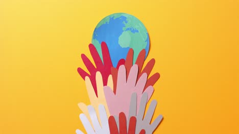 close up of hands together with globe made of colourful paper on yellow background with copy space