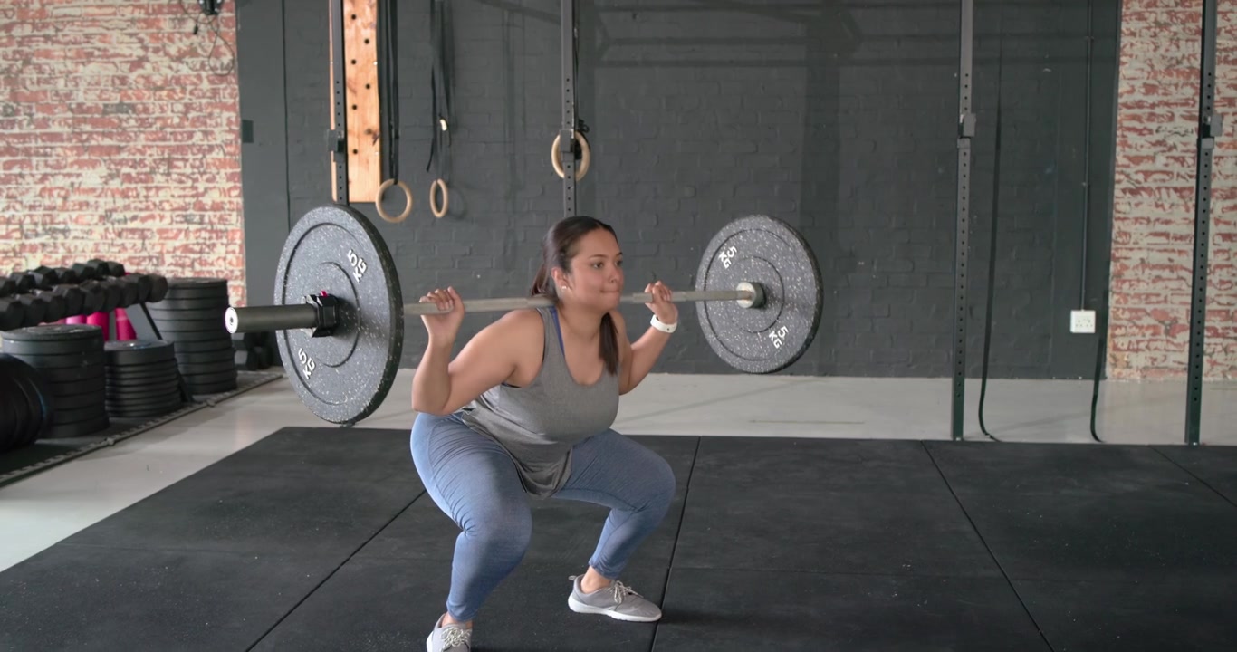 Atleta donna asiatica che afferra un bilanciere, facendo un passo indietro e accovacciandosi profondamente per la potenza in palestra.