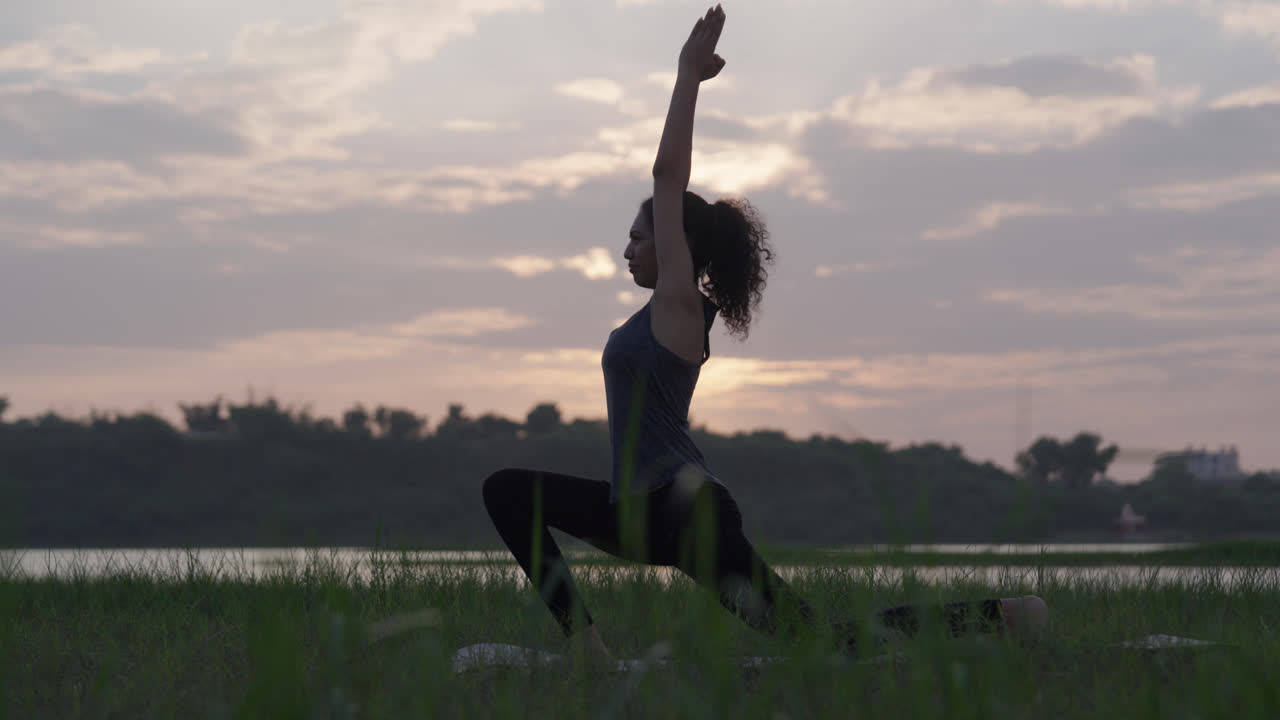 Una donna pratica yoga in posizione di affondo basso all'alba vicino a un fiume calmo con campi verdi