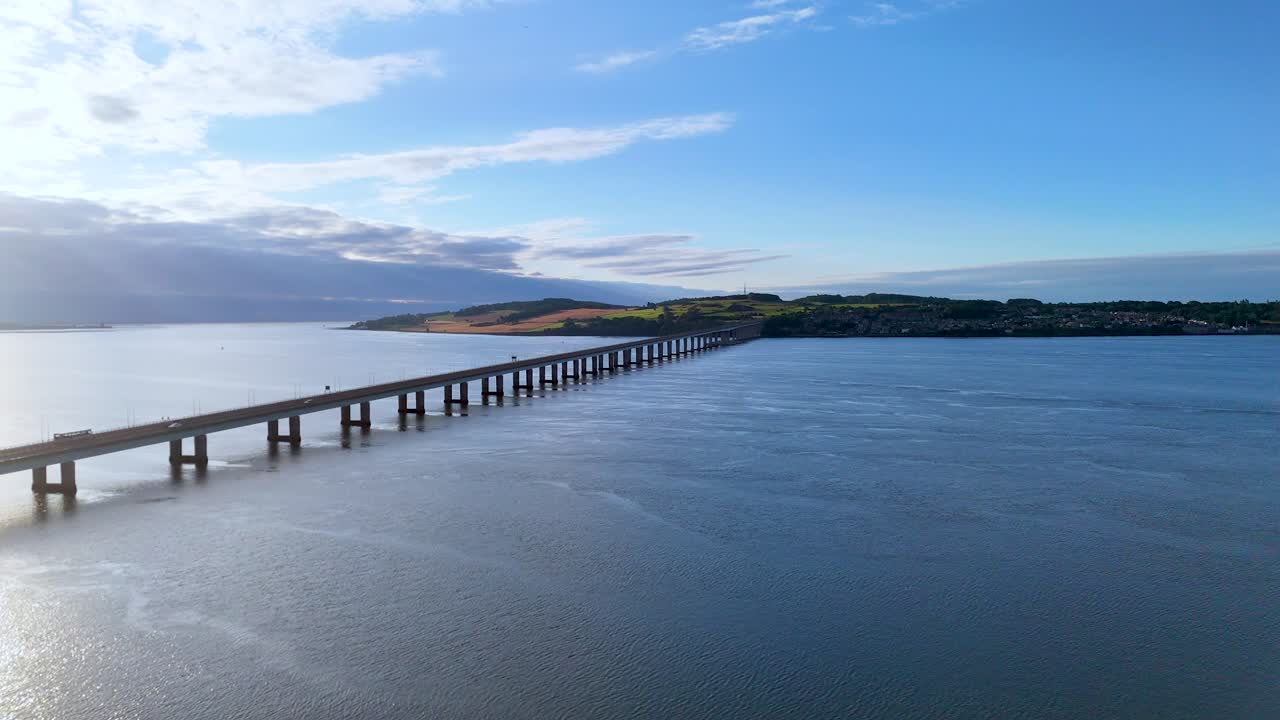 Aerial footage captures a long bridge spanning a wide river or estuary under bright daylight, with smooth camera movement and expansive scenic views