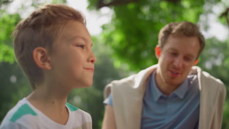 Munching-boy-laughing-talking-with-father-on-picnic.-Family-lunching-in-park.