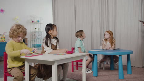 Female-Teacher-Giving-Colored-Pencils-To-Her-Pupils-Sitting-At-Desk-In-Montessori-School