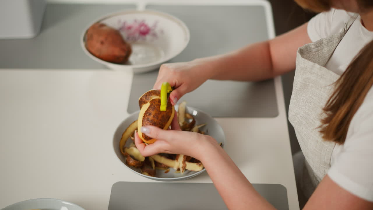 Close up of woman in apron peeling sweet potato over plate filled with skins while preparing food in kitchen, focused on hands and potato with smooth background featuring bowl with raw potatoes