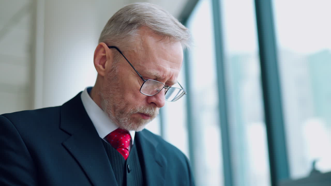 Portræt af en ældre forretningsmand ved vinduet. Moden mand med briller og i jakkesæt kigger ned, mens han arbejder på kontoret. Forretningskoncept.
