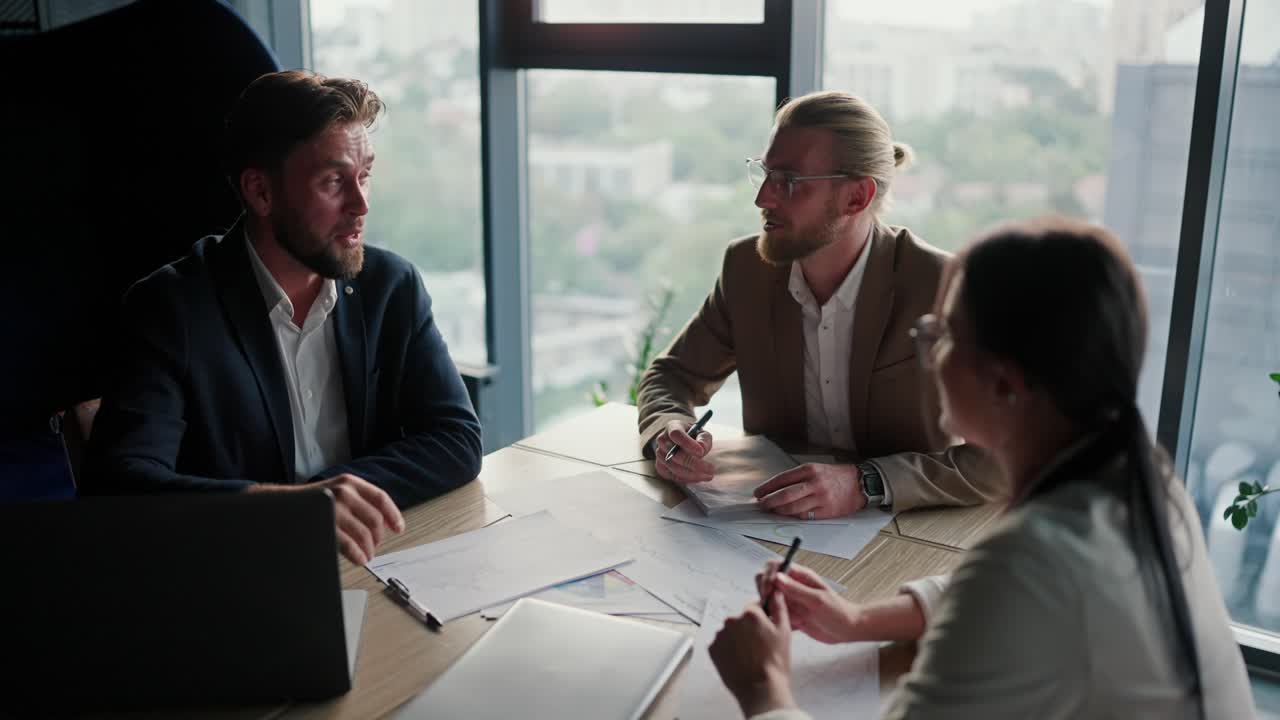 Three office workers, two guys and a girl in business suits, sit at the table and work with papers. Office workers writing on paper at a table in a modern office with a panoramic window