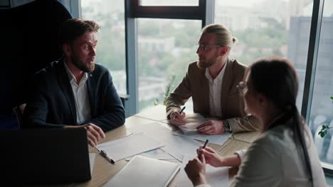 Three-office-workers,-two-guys-and-a-girl-in-business-suits,-sit-at-the-table-and-work-with-papers.-Office-workers-writing-on-paper-at-a-table-in-a-modern-office-with-a-panoramic-window