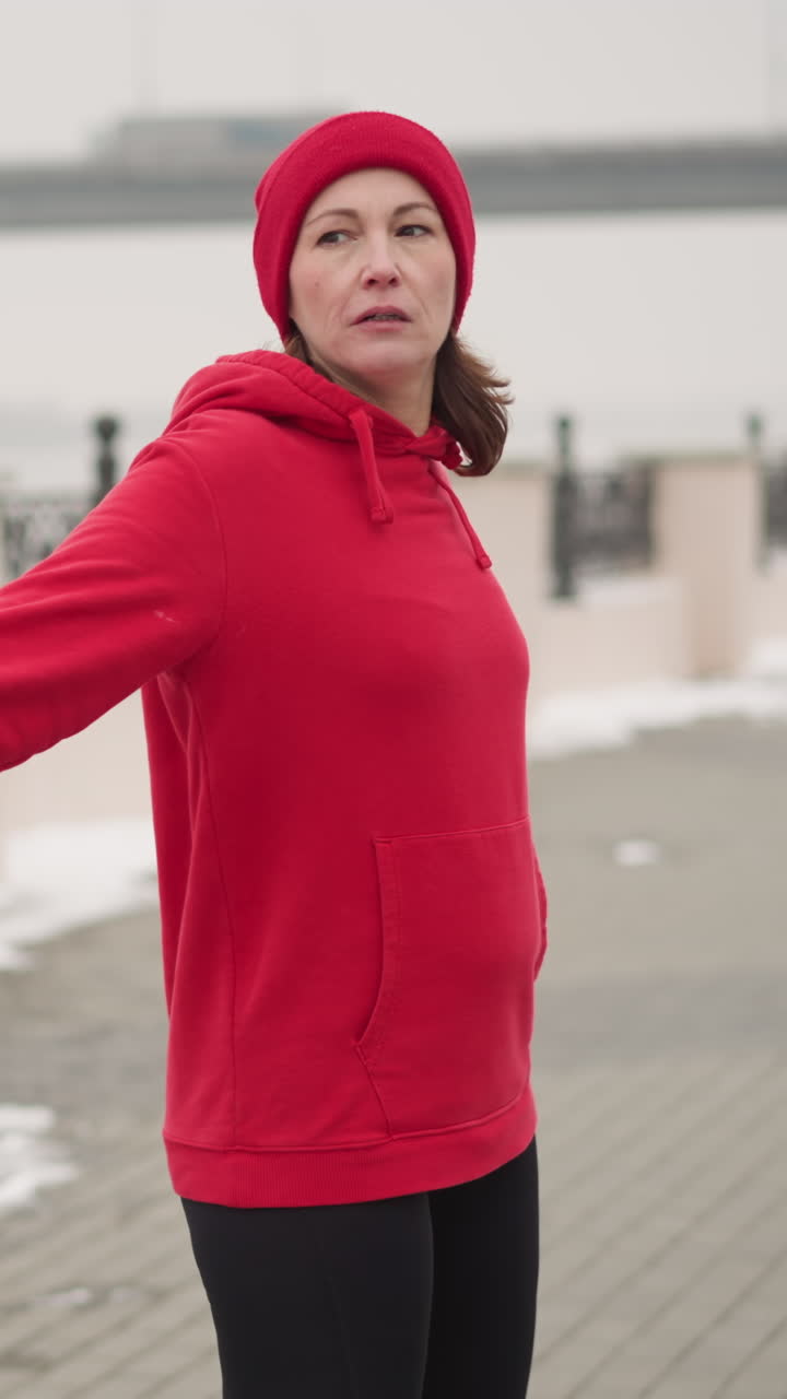 mujer con capucha roja y guantes realizando mano se extiende al aire libre en el camino cubierto de nieve con valla de metal decorativa, puente lejano, y coche en movimiento en atmósfera de niebla