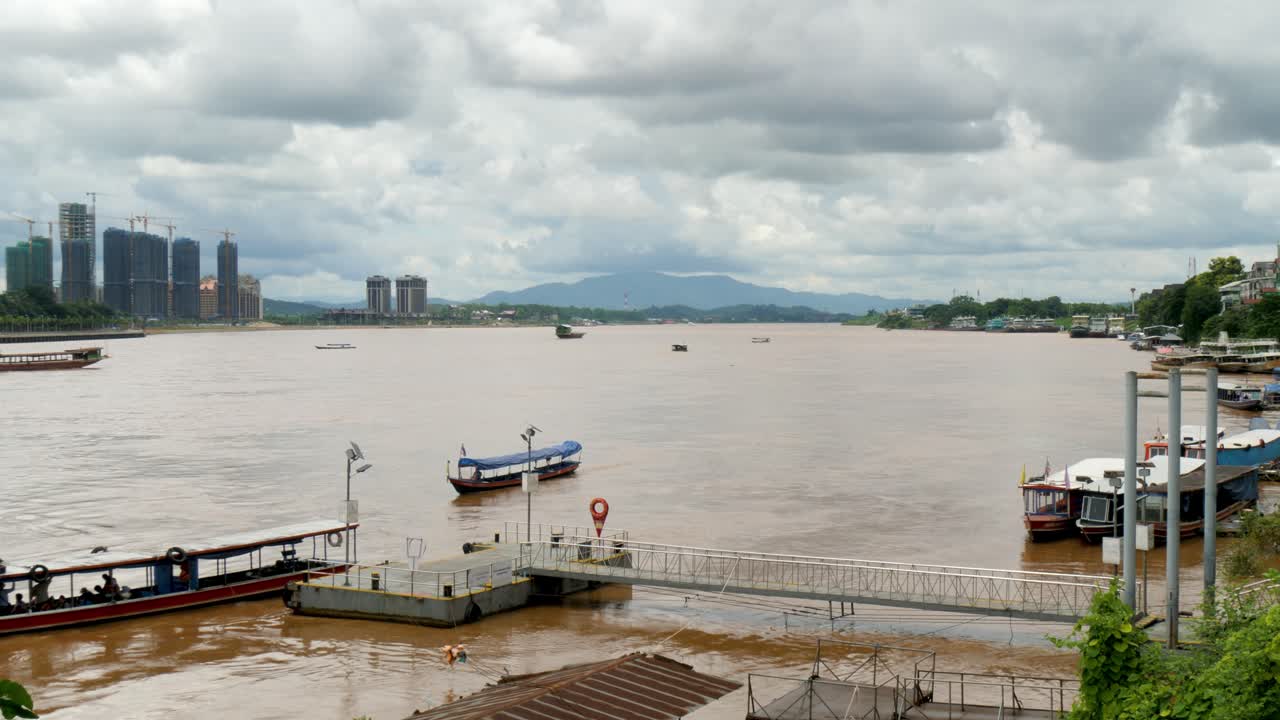 Ferry dock on the Mekong River in northern Thailand, with longtail boats and high-rise buildings under construction across the river in Laos under cloudy skies