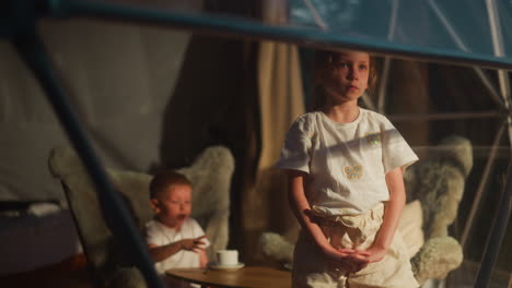 niña molesta mira por la ventana con el hermano menor en la habitación. niños pequeños aburridos cansados de jugar pasan el tiempo en casa. niños infelices en la cabaña de glamping