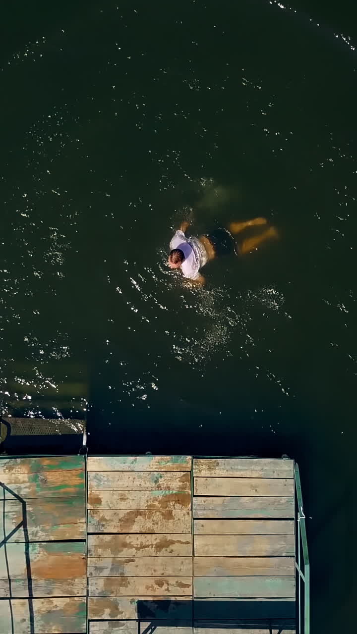Young man in white t-shirt falling down with his back into the water from the wooden bridge in summer. Male jumping in the river and swimming. Aerial view Vertical video