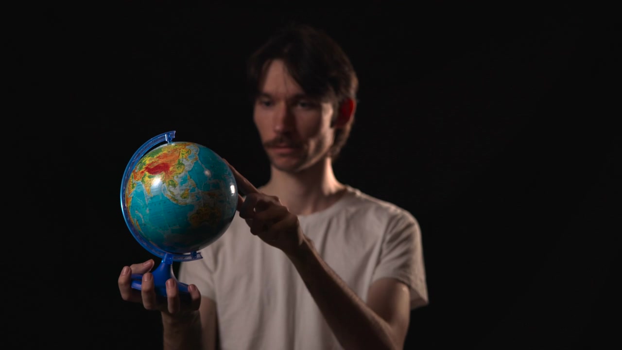 Un giovane uomo si staglia su uno sfondo nero, tenendo delicatamente e ruotando un mappamondo colorato con entrambe le mani, mentre una luce laterale soffusa illumina il suo viso e le braccia.