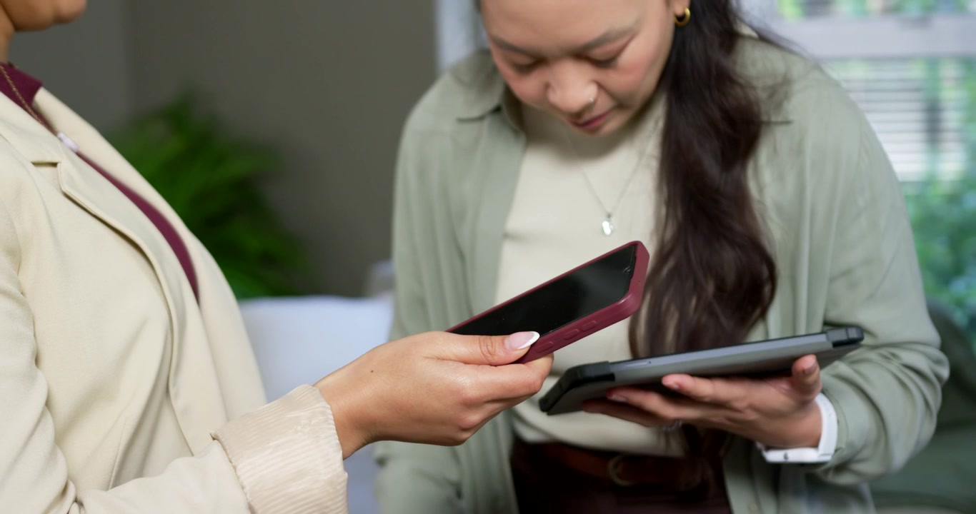 Diverse female coworkers comparing devices, left offering phone and right checking tablet on sofa