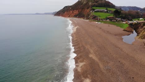 Toma-Aérea-Suave-De-4k-Bordeando-El-Borde-Del-Océano-Y-Vislumbrando-Las-Montañas-Y-Las-Colinas-Ondulantes-De-La-Playa-De-Branscombe