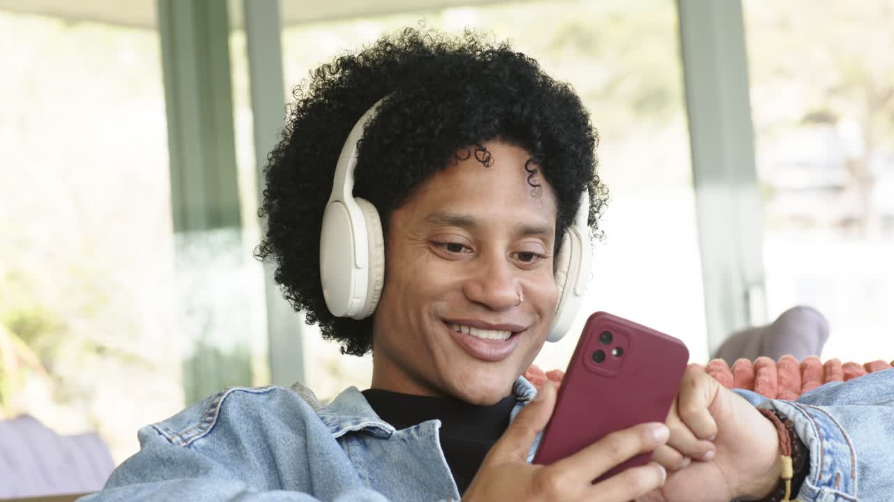 Uomo di mezza età con cuffie e braccialetti che sorride mentre scorre sul suo smartphone rosso seduto sul divano vicino alla porta a vetri