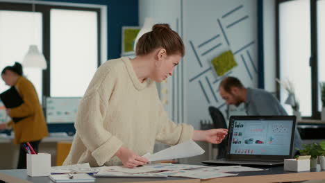 busy startup employee looking at papers with business data comparing numbers with laptop screen