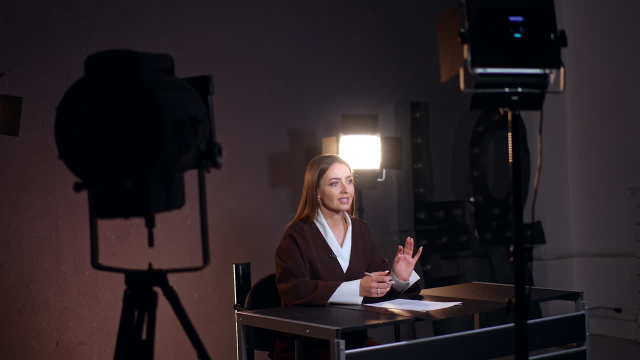 Beautiful confident Caucasian lady sits at desk talking to camera. Soffits stand around the female reporter. Studio footage.