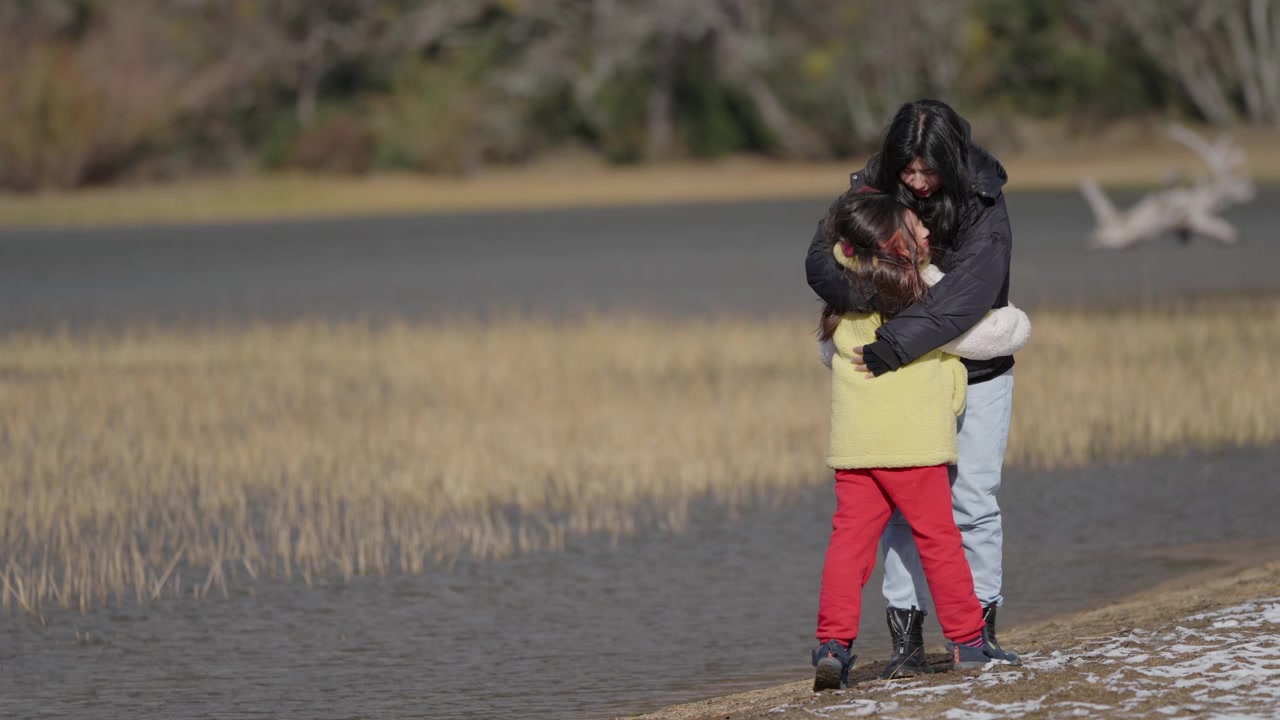 A mother and daughter exchange a heartfelt hug by a lakeside during winter. The scene captures a tender moment of connection. Perfect for family or lifestyle content.
