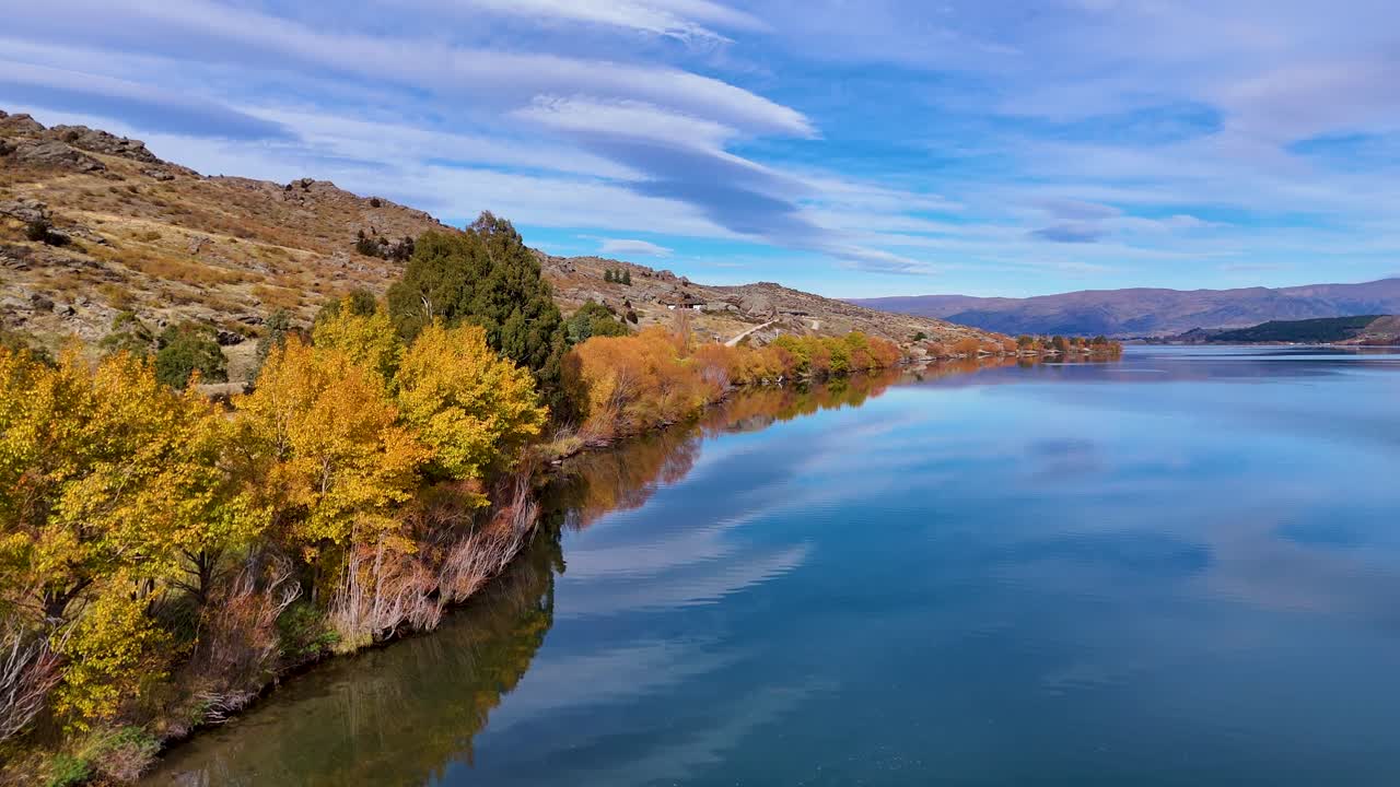Drone footage captures Lake Dunstan's serene waters and vibrant autumn foliage under a clear sky in Cromwell, New Zealand