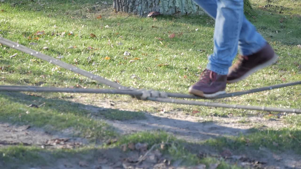 Man's legs balancing on rope in a park practicing slacklining. Walks forward then backwards. Woman's legs join on the other rope across. both drop down.