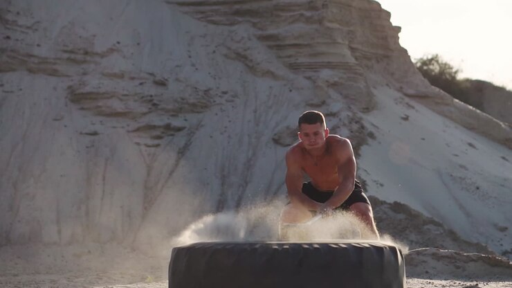 Un atleta strongman muscoloso colpisce una gigantesca ruota con un martello in montagna sabbiosa al rallentatore al tramonto. La polvere dalla ruota si solleva