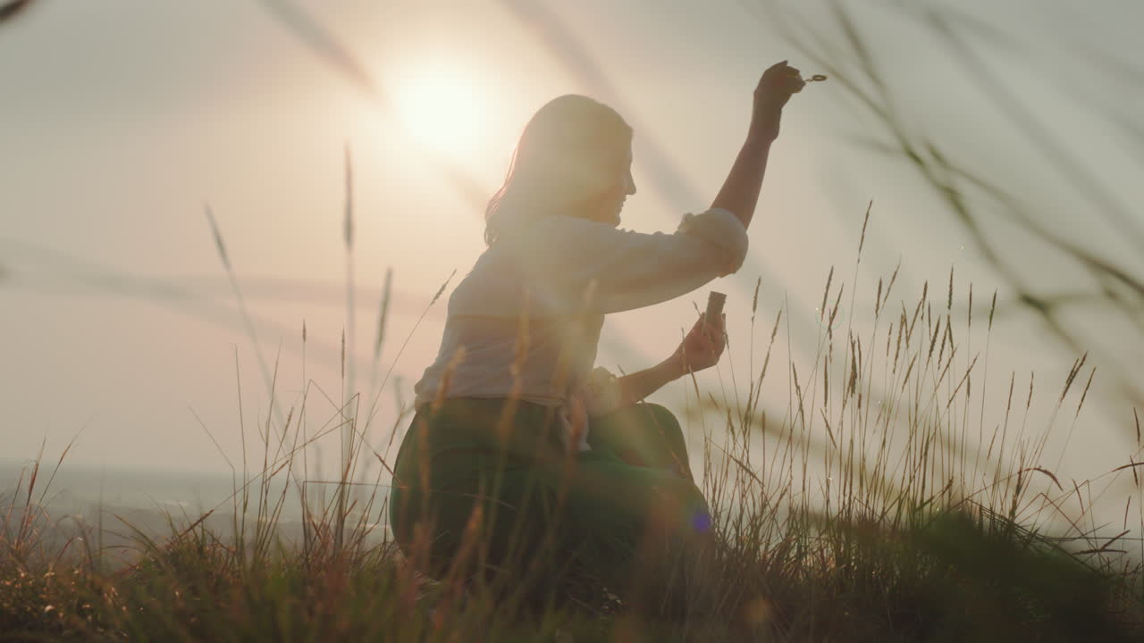 Silhouetted woman crouching in tall grass blows bubbles into golden sky during sunset, sunlight creating lens flare and soft atmospheric tones as bubbles drift through warm tranquil landscape