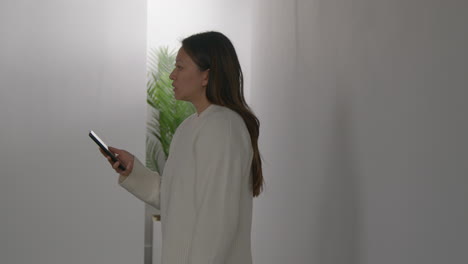 stressed or anxious woman leaning against wall at home reacting to internet or social media news message or story on mobile phone