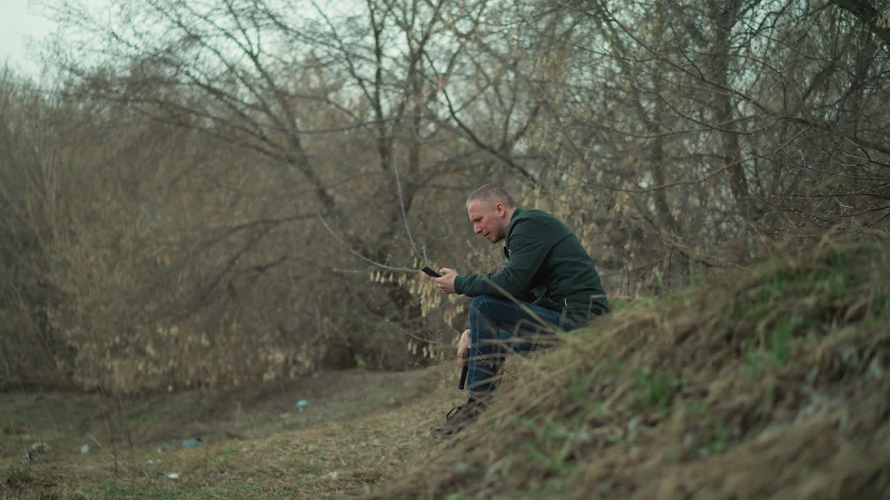 близкий вид человека, сидящего на травяном склоне холма с голыми деревьями на заднем плане, с пистолетом в руках и задумчиво глядя вниз, когда он управляет своим телефоном