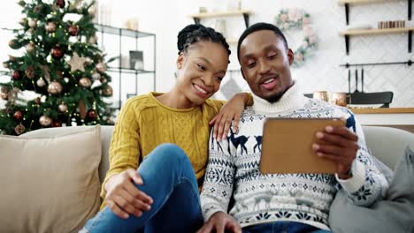 Portrait-Of-Happy-Family-Sitting-On-Sofa-In-Modern-Room-With-Decorated-Glowing-Christmas-Tree-And-Speaking-On-Video-Call-On-Tablet-At-Home-Saying-Holiday-Greetings-To-Friends