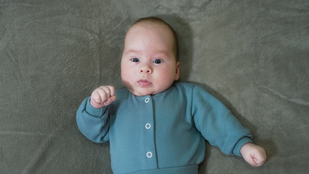 Little baby looks surprisingly into the camera. Beautiful kid in blue sweater lying on the grey soft plaid. Close up.