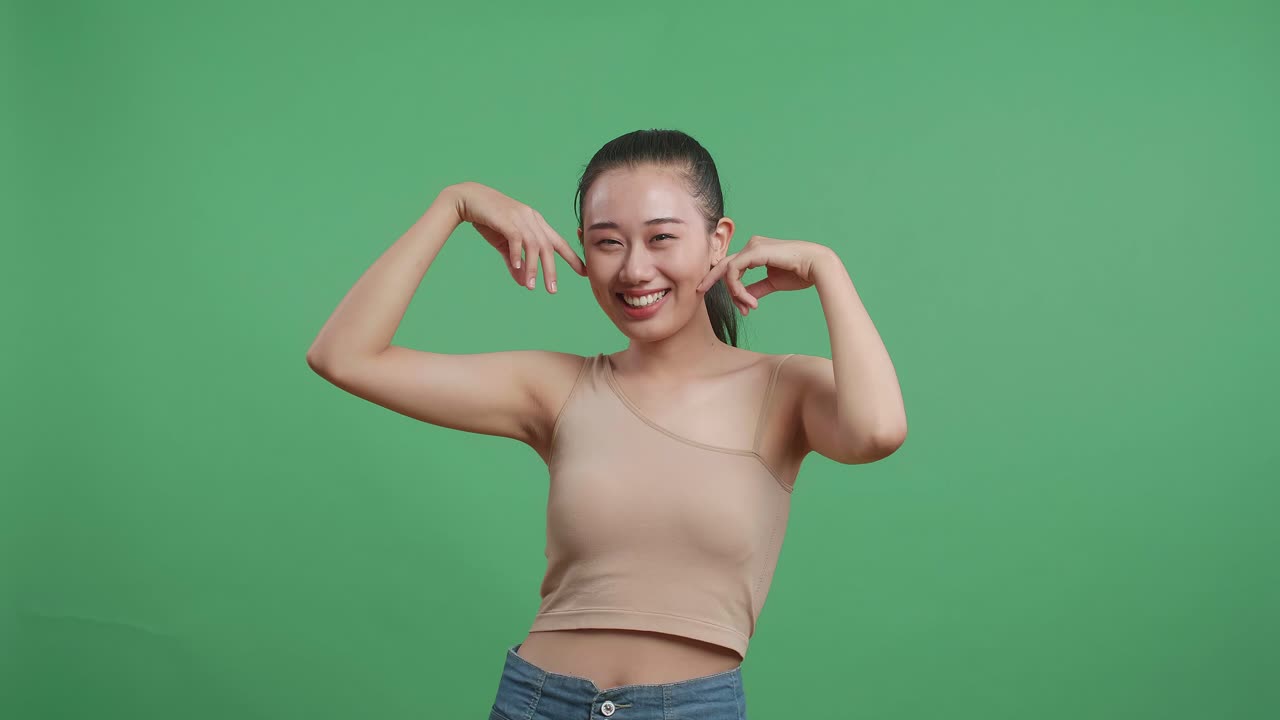 아름다운 아시아 여성이 인스 손가락으로 얼굴을 만지고 스튜디오의 녹색 화면 배경에서 카메라를 보고 미소 짓고 있습니다.