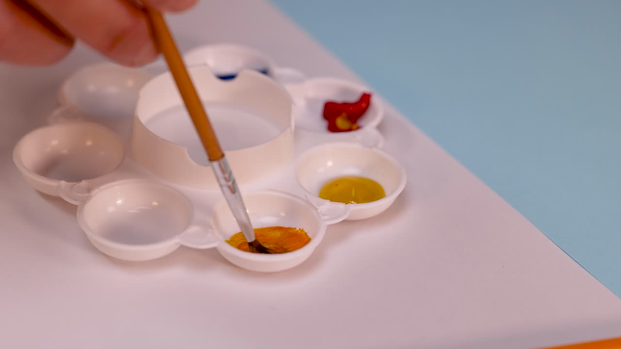 Close-up of hand mixing orange paint in palette using brush, soft lighting, steady overhead shot