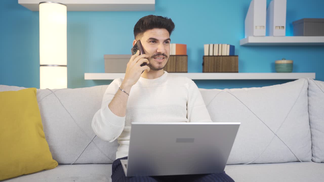 hombre riendo en casa hablando por teléfono y usando computadora portátil.
